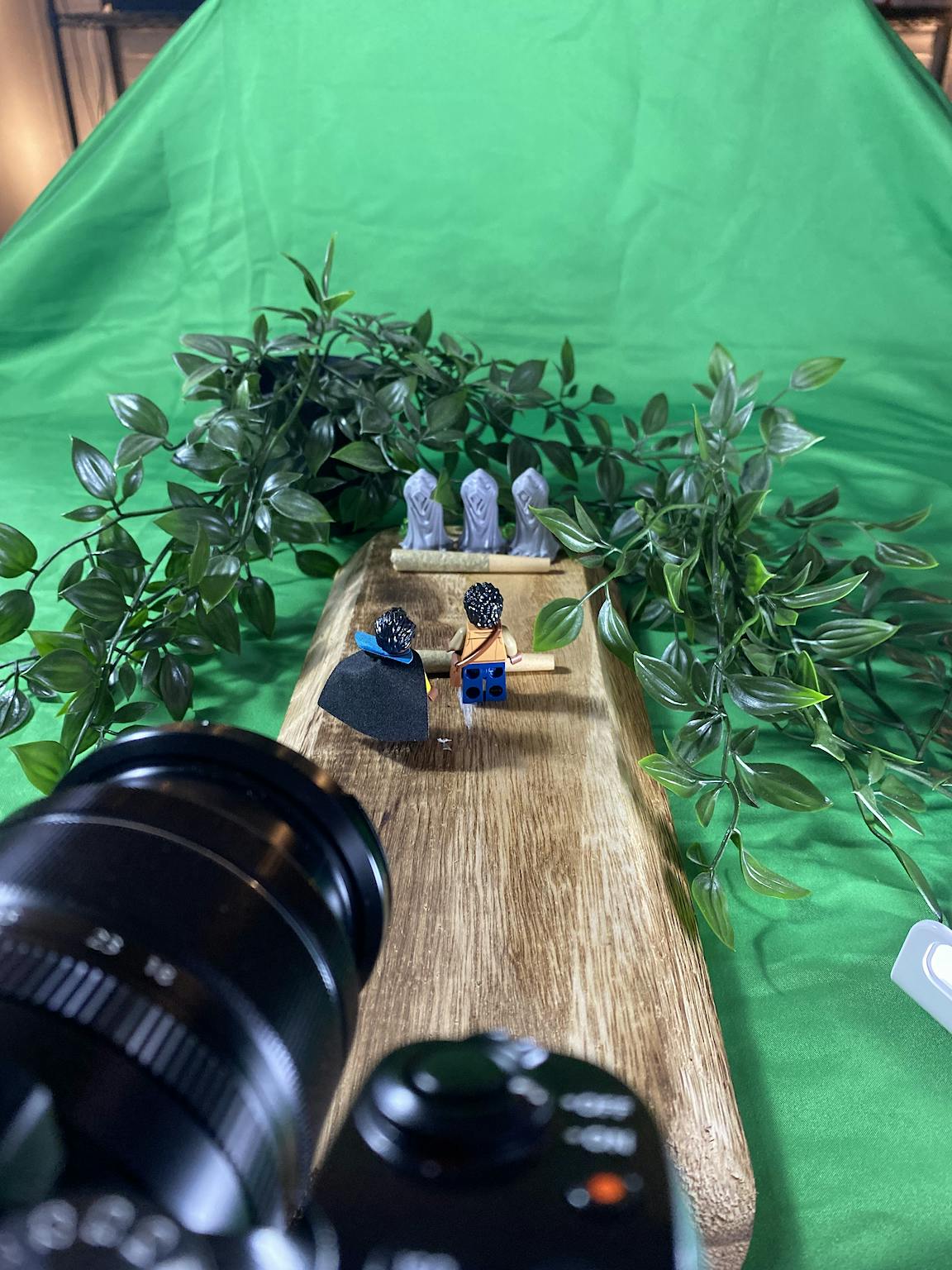 A camera is pointed at Lego characters in front of a leafy scene.